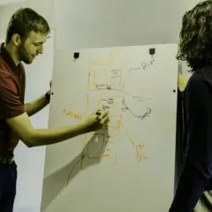 Dwóch mężczyzn planuje projekt na tablicy. Jeden pisze "content", drugi patrzy, jakby chciał uniknąć filistra.