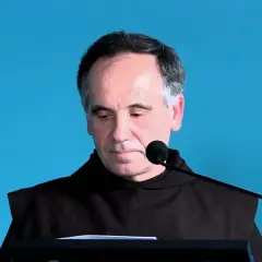 Mężczyzna w brązowym habicie, pochylony nad pulpitem, z mikrofonem przed twarzą. Jego wyraz twarzy sugeruje głębokie zamyślenie, być może związane z mistycyzmem.