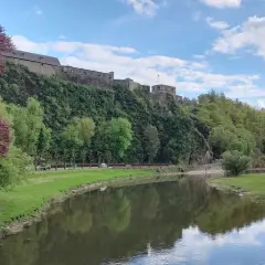 Zamek na wzgórzu w Ardenach, nad spokojną rzeką, otoczony zielenią i błękitnym niebem z chmurami.