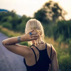 Blondynka z pierścionkiem w kształcie kwiatka, jak mały szelma, poprawia włosy na tle drogi i zieleni.
