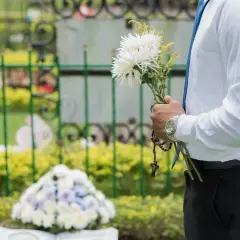 Mężczyzna w białej koszuli i niebieskim krawacie trzyma bukiet białych kwiatów i różaniec. W tle cmentarz, gdzie spoczywaj w pokoju.