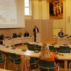 Ludzie siedzą przy okrągłym stole w sali konferencyjnej, słuchając prelegenta. To obraz ludzkiej egzystencji w przestrzeni wspólnego dyskursu.