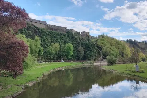 Zamek na wzgórzu w Ardenach, nad spokojną rzeką, otoczony zielenią i błękitnym niebem z chmurami.