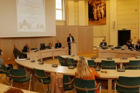 Ludzie siedzą przy okrągłym stole w sali konferencyjnej, słuchając prelegenta. To obraz ludzkiej egzystencji w przestrzeni wspólnego dyskursu.