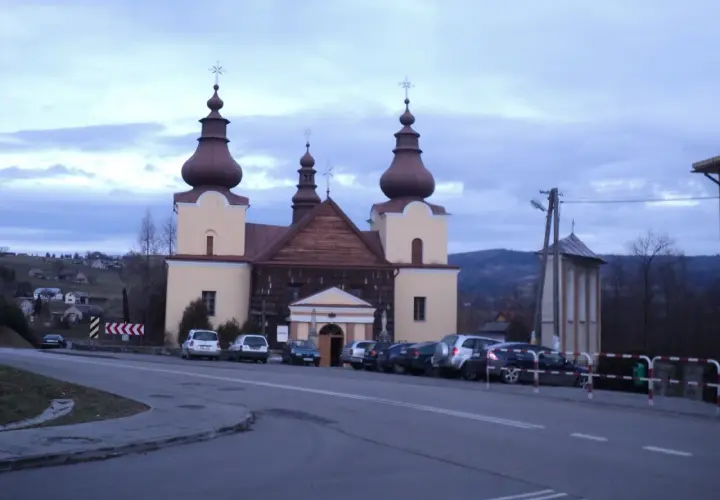 Cerkiew łemkowska z charakterystycznymi kopułami, otoczona samochodami, na tle pagórkowatego krajobrazu.