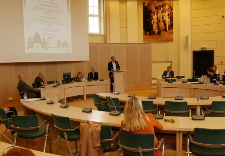 Ludzie siedzą przy okrągłym stole w sali konferencyjnej, słuchając prelegenta. To obraz ludzkiej egzystencji w przestrzeni wspólnego dyskursu.