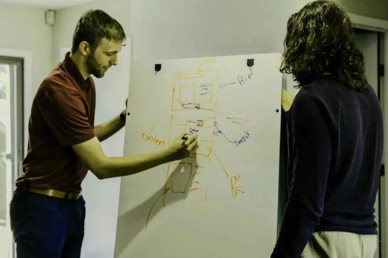Dwóch mężczyzn planuje projekt na tablicy. Jeden pisze "content", drugi patrzy, jakby chciał uniknąć filistra.