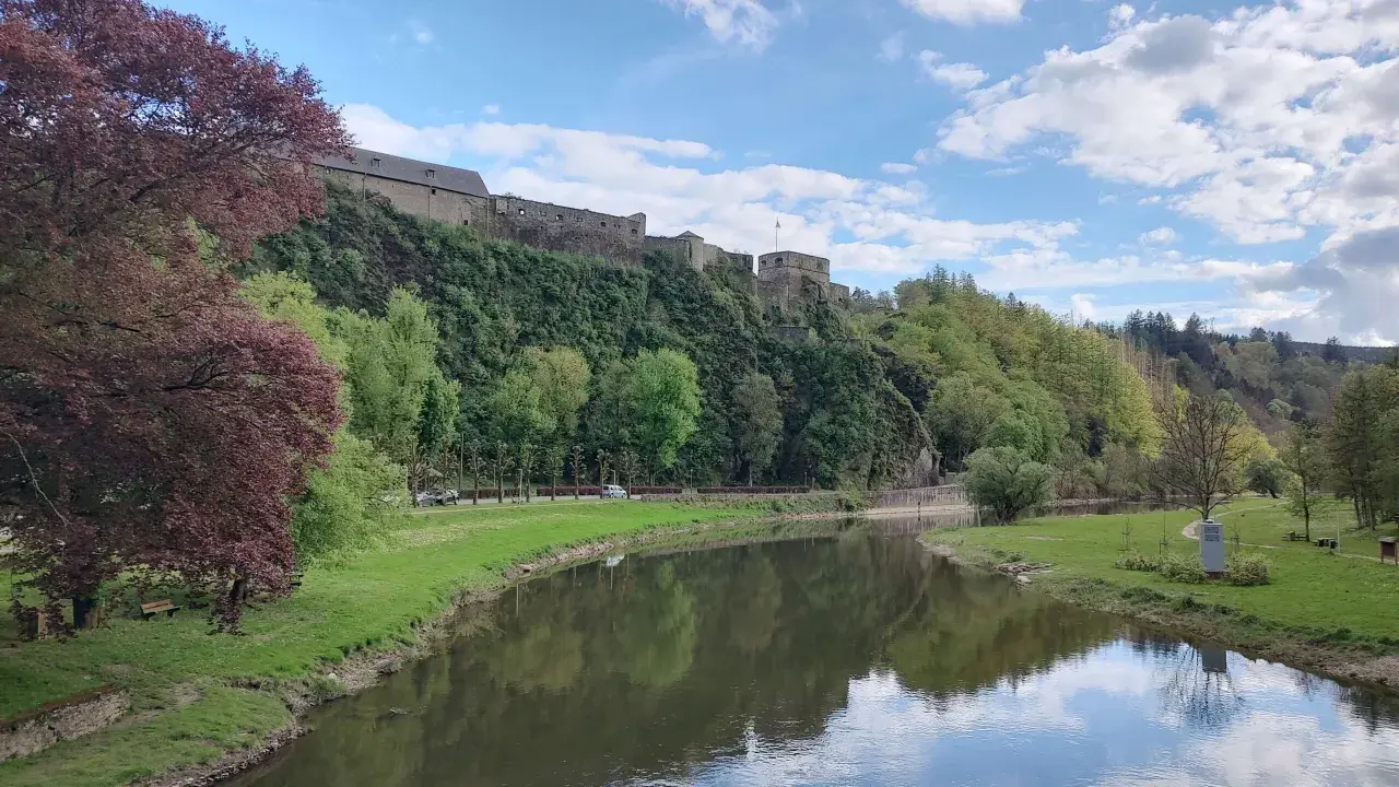 Zamek na wzgórzu w Ardenach, nad spokojną rzeką, otoczony zielenią i błękitnym niebem z chmurami.