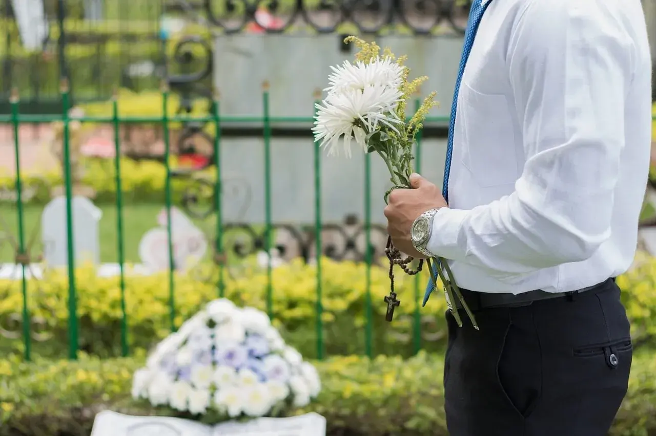 Mężczyzna w białej koszuli i niebieskim krawacie trzyma bukiet białych kwiatów i różaniec. W tle cmentarz, gdzie spoczywaj w pokoju.