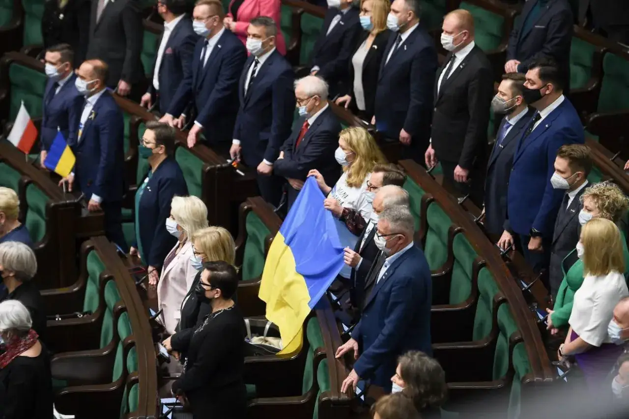 Ludzie w sali sejmowej z flagą Ukrainy, wyrażając solidarność. Wszyscy stoją, w geście aklamacji i wsparcia.